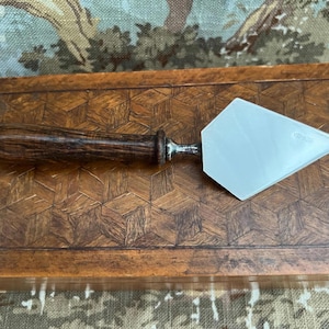 May include: A cake server with a wooden handle and a silver-colored blade. The handle is dark brown and the blade is a light silver color. The server is resting on a wooden surface with a geometric pattern.