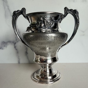 May include: A silver-toned metal trophy cup with two ornate handles. The front features embossed floral details and engraved text reading INDUSTRIAL TRUST COMPANY TENNIS TOURNAMENT and Runner Up. The cup has a wide rim and a circular pedestal base.