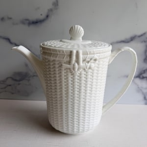 May include: A white ceramic teapot featuring a textured basket-weave pattern across the body and lid. The lid is topped with a small shell-shaped finial, and the side of the pot displays a decorative molded knot detail near the rim.