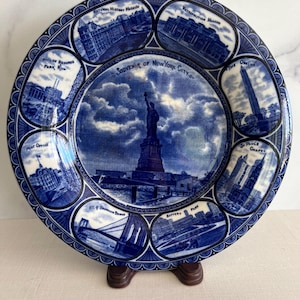 May include: A blue and white ceramic souvenir plate featuring the Statue of Liberty in the center. The plate is decorated with images of New York City landmarks, including the Metropolitan Museum and Brooklyn Bridge.