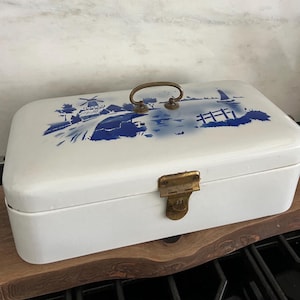 May include: White rectangular bread box with a blue and white landscape design on the lid. The design features a windmill, water, and a sailboat. It has a brass handle and clasp.