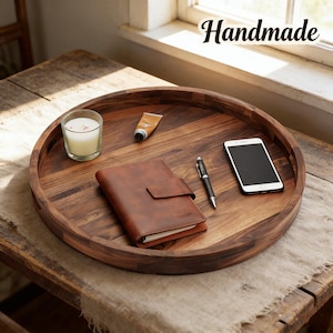 May include: A round, dark wood serving tray with a variety of items on it, including a brown leather journal, a pen, a smartphone, a candle, and a tube of cream. The word "Handmade" is written in cursive.