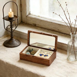 May include: A wooden jewelry box with a mirrored lid, open to reveal compartments with rings and a pineapple-shaped trinket. A lit candle sits in a decorative metal holder. A glass vase with branches is also present.