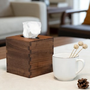 May include: A wooden tissue box with dovetail joints, holding white tissues. Beside it, a white mug with a black rim and a few dried flowers. A pinecone rests nearby. The items are on a white surface.