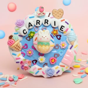 May include: A round blue decorative plaque featuring the name CARRIE in white letter beads. The border is decorated with white icing, colorful sprinkles, a central white bunny figure, miniature ice cream cones, flowers, and a heart-shaped cookie.