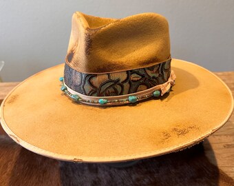 May include: A tan felt cowboy hat with a wide brim and a leather band decorated with turquoise beads and a floral pattern.