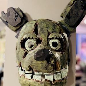May include: A green and brown mask with large eye sockets, a wide mouth, and long, upright ears. The mask has a distressed, textured appearance, with visible cracks and a worn aesthetic. The mask is mounted on a light-colored wall.