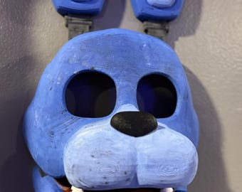 May include: A blue bunny mask with large, dark eye sockets, a black nose, and a wide open mouth revealing white teeth and a red interior. The mask has large, upright ears and is likely a costume or decorative item.