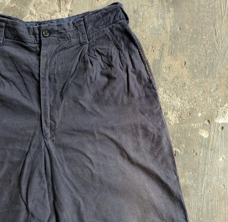 May include: Close-up of dark navy blue shorts. The shorts have a button closure and pleats at the waist. The fabric appears to be a soft, woven material. The shorts are slightly wrinkled, suggesting they are made of a natural fiber.