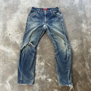 May include: Faded blue denim jeans with distressed details, including rips at the knees and frayed hems. The jeans have a classic five-pocket design, a button and zipper closure, and a red label at the waistband.