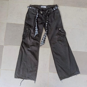 May include: Dark brown cargo pants with a wide leg design. The pants feature two front pockets, two side cargo pockets, and a decorative belt with a skull pattern. The pants are made of a durable fabric.