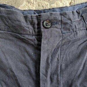 May include: Close-up of navy blue pants with a button closure. The pants have a button and a buttonhole, with visible stitching details. The fabric appears to be a woven material.
