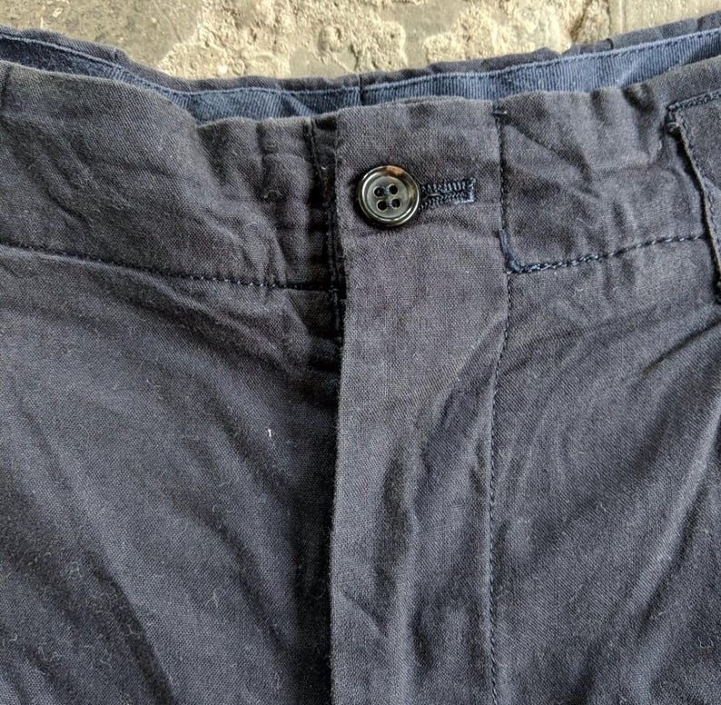 May include: Close-up of navy blue pants with a button closure. The pants have a button and a buttonhole, with visible stitching details. The fabric appears to be a woven material.