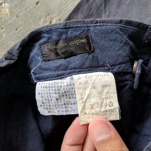 May include: Close-up of a navy blue garment tag with the brand name "COMME des GARCONS" and Japanese text. The tag includes care instructions and a size M label. The fabric is 100% cotton.