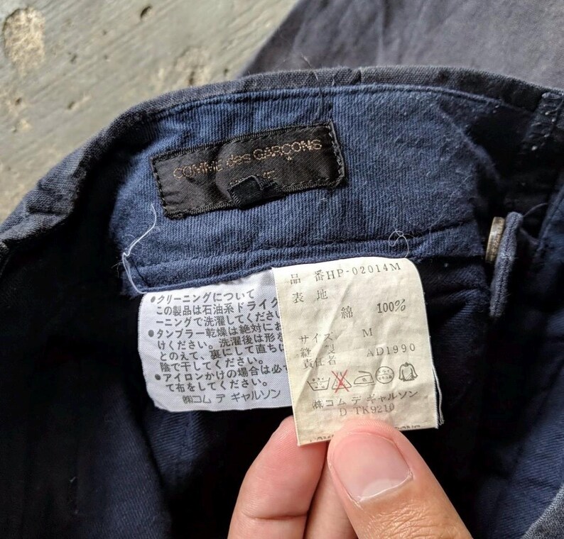 May include: Close-up of a navy blue garment tag with the brand name "COMME des GARCONS" and Japanese text. The tag includes care instructions and a size M label. The fabric is 100% cotton.