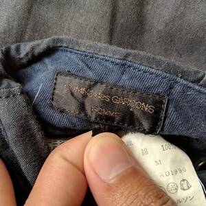 May include: Close-up of a dark gray shirt collar with a navy blue interior. A black label reads "COMME des GARCONS HOMME" in gold lettering. A white tag with Japanese text and "100%" is also visible.