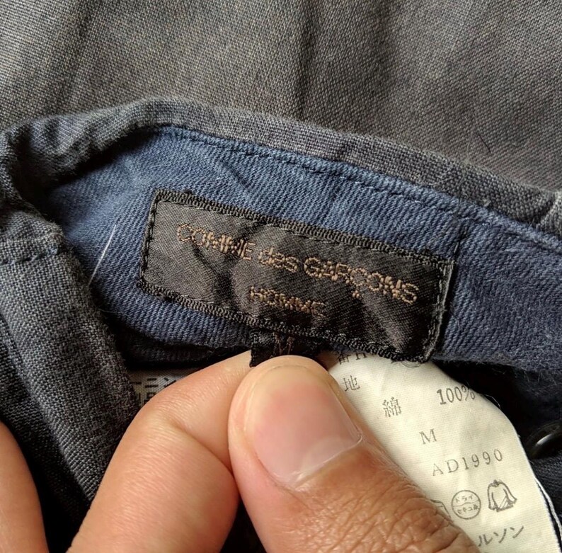 May include: Close-up of a dark gray shirt collar with a navy blue interior. A black label reads "COMME des GARCONS HOMME" in gold lettering. A white tag with Japanese text and "100%" is also visible.