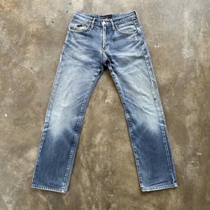 May include: Faded blue denim jeans with a classic five-pocket design. The jeans have a straight leg cut and a button and zip closure. The denim has a worn appearance with lighter areas on the thighs and knees, and darker areas on the lower legs.