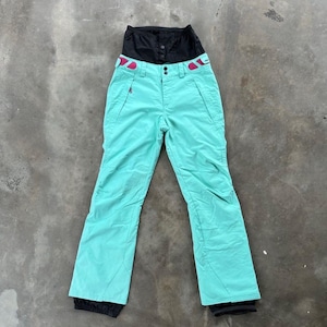 May include: Mint green ski pants featuring a high-waisted black integrated gaiter, a front zipper closure with two buttons, and two small pink accents on the waistband. The pants have two side pockets and black trim at the bottom leg openings.