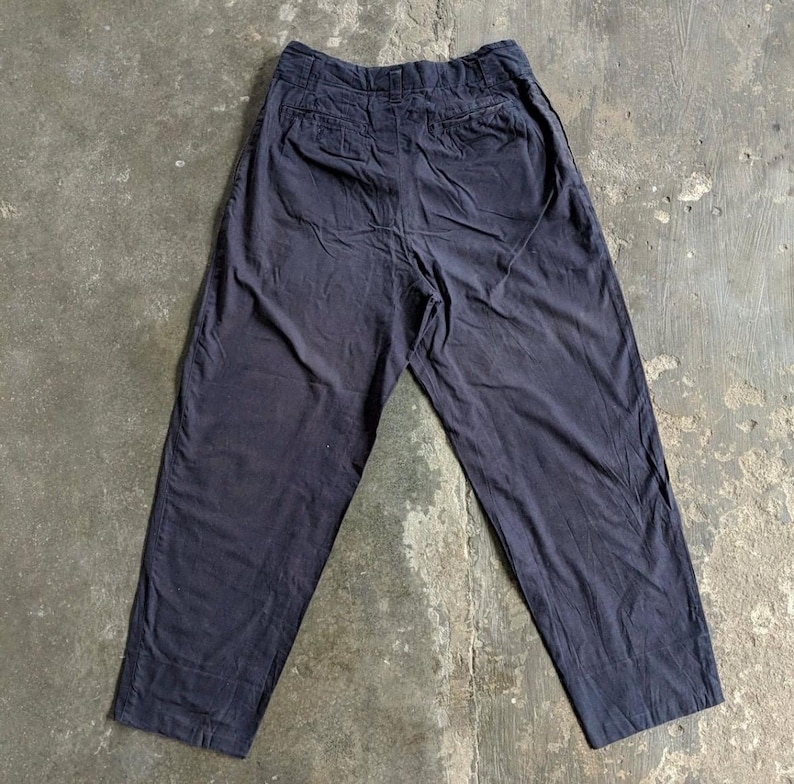 May include: Dark navy blue pants with a high waist and two back pockets. The pants are made of a soft, slightly wrinkled fabric. The legs are straight and wide, giving a relaxed fit. The pants are laid flat on a concrete surface.