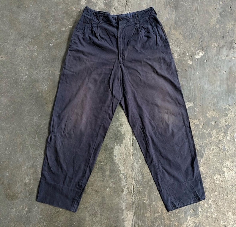 May include: Dark navy blue pants with a high waist and pleated front. The pants have a relaxed fit and a button closure. The fabric appears worn, with lighter areas and subtle fading, suggesting a vintage or well-loved item.