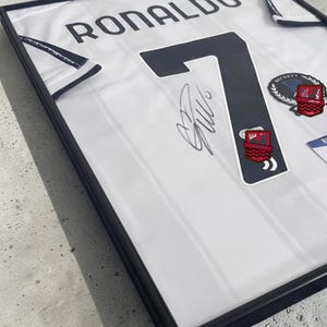 Pode incluir: Uma camisola desportiva branca com acabamentos pretos, emoldurada numa moldura preta. A camisola apresenta o nome RONALDO e o número 7 a preto, com uma assinatura a tinta escura ao lado do número. Um selo de autenticação Beckett é visível.