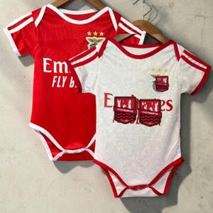Op de afbeelding: Twee babyrompertjes op kleerhangers. De ene is rood met witte randen, voorzien van de tekst Emirates FLY BETTER en een embleem met drie sterren. De andere is wit met rode randen, voorzien van de tekst Emirates, een embleem en twee COZYBERRYW-schildpatches.