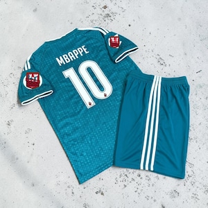Puede incluir: Un conjunto de fútbol color verde azulado que incluye una camiseta y pantalones cortos a juego. La camiseta muestra el nombre MBAPPE y el número 10 en blanco en la espalda, con rayas blancas en las mangas y un pequeño escudo en cada hombro.