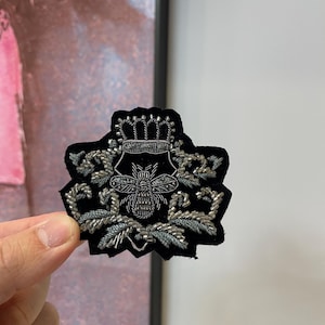 May include: Black velvet patch with a silver embroidered bee design, crowned and surrounded by floral accents. The patch is held up by a hand, showcasing the intricate details of the embroidery. The patch is a decorative item.