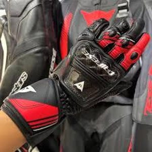 Puede incluir: Guantes de moto negros y rojos con la marca "DAINESE" visible. Los guantes tienen un protector de nudillos de fibra de carbono y una correa de velcro en la mu&ntilde;eca. El dise&ntilde;o incluye detalles en blanco y un logotipo triangular.
