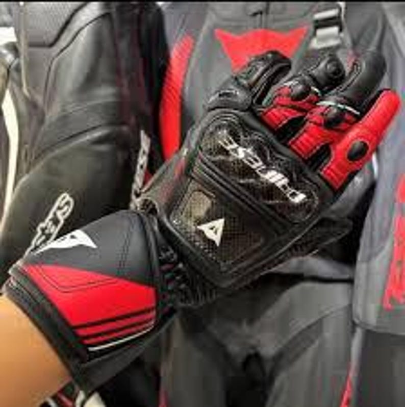 Puede incluir: Guantes de moto negros y rojos con la marca "DAINESE" visible. Los guantes tienen un protector de nudillos de fibra de carbono y una correa de velcro en la mu&ntilde;eca. El dise&ntilde;o incluye detalles en blanco y un logotipo triangular.