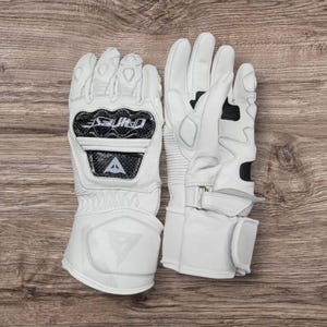 May include: A pair of white motorcycle gloves with black accents. The gloves feature a carbon fiber knuckle protector and a wrist strap. The brand name "DAINESE" is visible on the gloves. The gloves are made of leather and have a padded design.