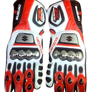 May include: Pair of motorcycle gloves in white, red, and black. The gloves feature the Suzuki logo and GSX branding. The design includes protective padding on the knuckles and fingers, with a combination of leather and synthetic materials.
