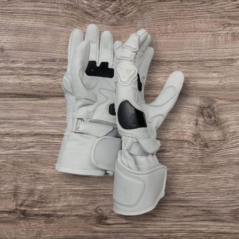 May include: A pair of white motorcycle gloves with protective padding on the knuckles and wrist. The gloves have black accents and a secure strap closure. The gloves are made of leather and are designed for safety and comfort.