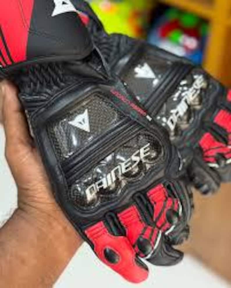 Puede incluir: Guantes de moto negros y rojos con la marca "DAINESE". Los guantes tienen nudillos reforzados, dise&ntilde;o de fibra de carbono y una combinaci&oacute;n de cuero y materiales sint&eacute;ticos. Dise&ntilde;ados para protecci&oacute;n y rendimiento.