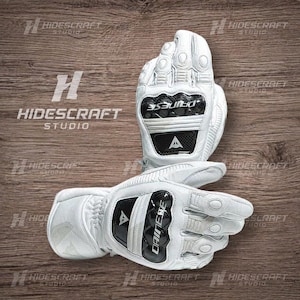 May include: White motorcycle gloves with black accents and the brand name "DAINESE" visible. The gloves have protective features on the knuckles and palms. The gloves are displayed on a wooden surface.