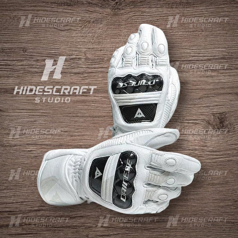 May include: White motorcycle gloves with black accents and the brand name "DAINESE" visible. The gloves have protective features on the knuckles and palms. The gloves are displayed on a wooden surface.
