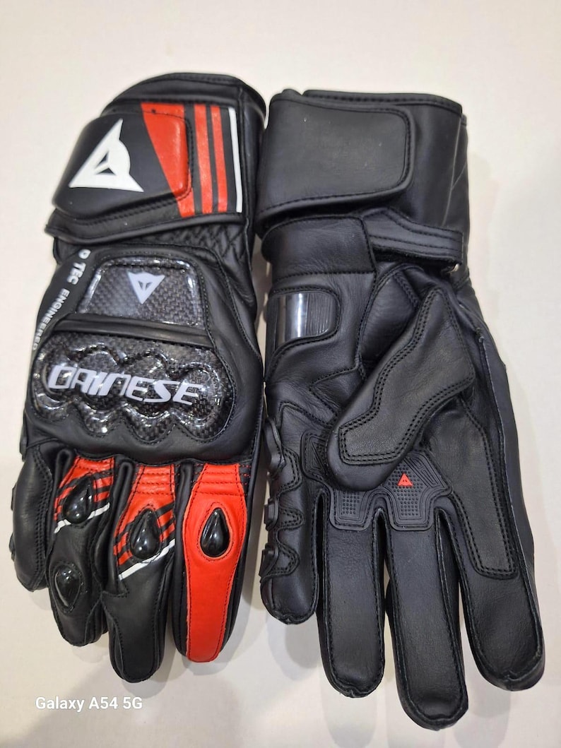 Puede incluir: Par de guantes de moto negros con detalles rojos y blancos. Los guantes cuentan con un protector de nudillos de fibra de carbono y la marca "DAINESE". El dise&ntilde;o incluye una correa de mu&ntilde;eca y una palma reforzada.