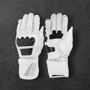 May include: White motorcycle gloves with black detailing. The gloves display the brand name "DAINESE" in black lettering on the knuckles and wrist. The gloves are made of leather and have protective padding for safety.