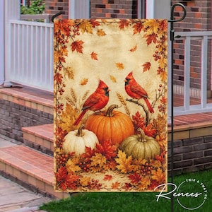 May include: A rectangular garden flag featuring two red cardinal birds perched near three pumpkins of varying sizes. The scene is framed by autumn foliage in shades of orange, yellow, and red, set against a textured, light beige background.
