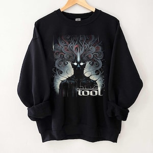 Puede incluir: Sudadera negra con un diseño gráfico que presenta una figura silueteada y la palabra "tool" en blanco. El diseño incluye elementos arremolinados rojos, azules y grises, y una luz brillante que emana de los ojos de la figura.