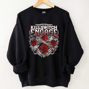 Può includere: Felpa nera con la scritta "Killswitch Engage" in rosso e bianco, circondata da rose rosse e un design decorativo. La felpa è appesa a una gruccia di legno.