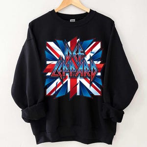 Könnte beinhalten: Schwarzes Sweatshirt mit einem grafischen Design der britischen Flagge in Rot, Weiß und Blau und dem Schriftzug "Def Leppard" in einer stilisierten Schriftart. Das Sweatshirt hängt an einem Holzkleiderbügel.