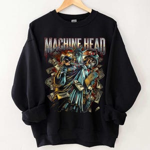 May include: Black sweatshirt featuring a graphic design with the text "MACHINE HEAD". The design includes an illustration of the Statue of Liberty holding a skull and a book, surrounded by money.