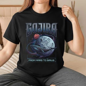 以下が含まれることがあります： 黒いTシャツ。バンド名「GOJIRA」が様式化されたフォントで描かれ、月と惑星を背景にクジラが泳いでいるグラフィックの上に配置されています。「FROM MARS TO SIRIUS」の文字が下にあります。