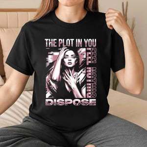 May include: Black t-shirt featuring a graphic of a woman with the text "THE PLOT IN YOU FEEL NOTHING DISPOSE" in pink and white. The woman is depicted with her hand on her head and chest.