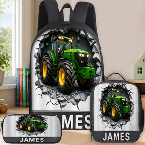 Puede incluir: Un conjunto de mochila, bolsa de almuerzo y estuche negros con un gráfico de un tractor verde emergiendo de un muro de hormigón agrietado. Cada artículo muestra el nombre JAMES en letras mayúsculas blancas sobre una franja gris oscuro.