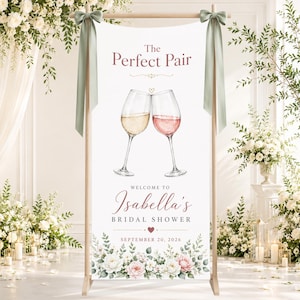 May include: A vertical banner on a wooden stand with sage green ribbons, featuring the text The Perfect Pair, Welcome to Isabella's Bridal Shower, and September 20, 2026. It displays two clinking wine glasses and a floral arrangement at the base.