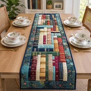 May include: A quilted table runner featuring a design of stacked books in various colors, including red, blue, and gold. The runner has a dark teal border with floral patterns. Tea cups and saucers are set on the table.