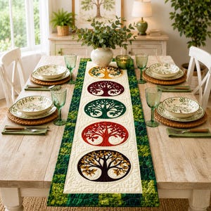 May include: A rectangular table runner featuring five circular embroidered tree motifs in yellow, maroon, green, red, and brown, set against a cream background with a green quilted border, displayed on a wooden dining table with place settings.